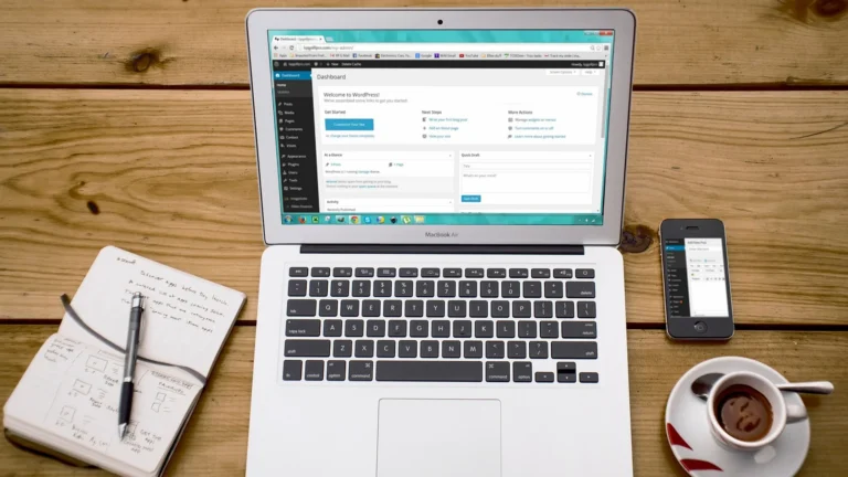 Tampilan dashboard WordPress pada layar laptop MacBook Air di atas meja kayu dengan buku catatan, ponsel, dan secangkir kopi.